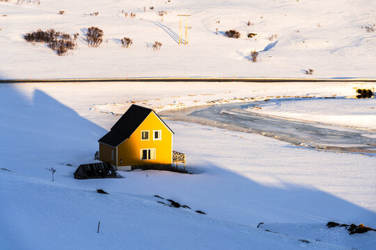 View of a striking yellow house basking under the sun's embrace amidst the vast white expanse of snow, creating a serene contrast, Sorvaer, Soroya island, Finnmark, Norway.