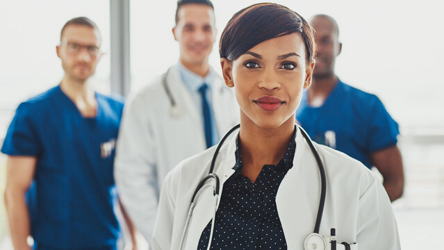 A black female doctor stands confidently in the foreground, looking directly into the lens. Behind her, a diverse medical team is visible, creating a sense of leadership and in a hospital setting.