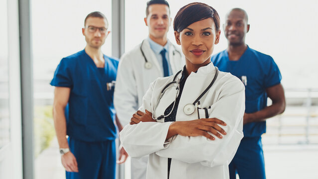 A black female doctor stands confidently in the foreground, looking directly into the lens. Behind her, a diverse medical team is visible, creating a sense of leadership and in a hospital setting.