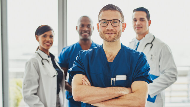 A smiling young doctor with glasses stands in the foreground, looking directly into the camera. Behind him, a diverse medical team is busy discussing a patient's case in the hospital hallway