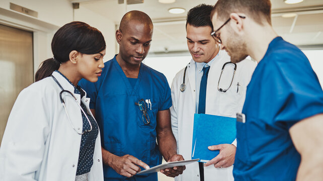 A multiracial team of four doctors in scrubs and white coats stands together, seriously reviewing medical papers in a hospital