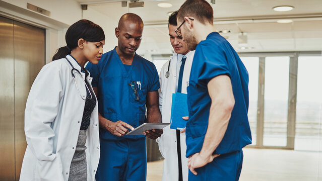 A multiracial team of four doctors in scrubs and white coats stands together, seriously reviewing medical papers in a hospital