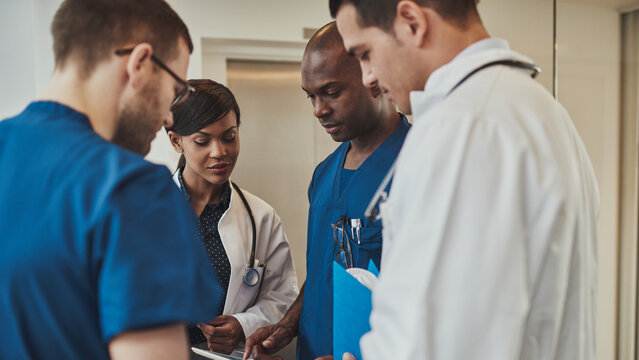 A diverse team of four doctors stands together in a hospital, deeply focused as they review medical documents. They all share a serious expression, reflecting the gravity of their discussion. 