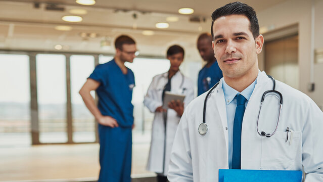 A male doctor stands in the foreground, looking directly into the camera with confidence. Behind him, a diverse medical team is gathered, highlighting a collaborative hospital environment