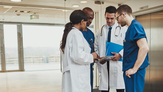 A multiracial team of doctors is gathered in a hospital hallway, reviewing some documents together.