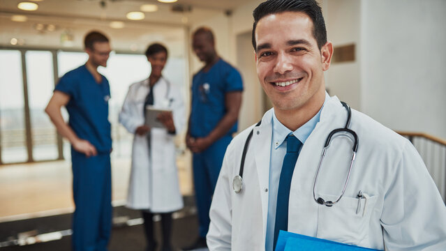 A smiling physician stands in a hospital hallway, clutching files under his arm and looking straight into the lens. In the background, his diverse medical team is discussing a patient's case.