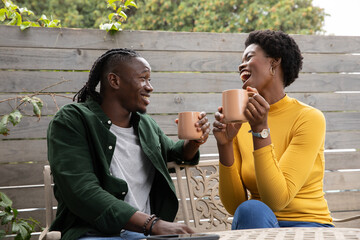 African american couple sitting on metal patio bench with ceramic mugs chatting with smartphone