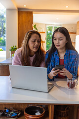 Asian mother and daughter checking smartphone and laptop screens at kitchen island with tea