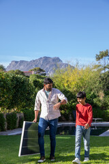 Indian father son walking on green lawn beside pool carrying solar panel with mountain backdrop