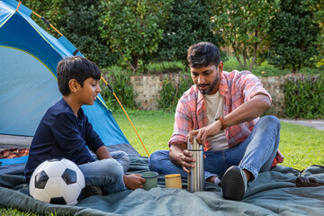 Indian father with son sitting cross-legged on lawn in front of blue tent, pouring from thermos © wavebreak3
