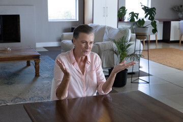 Obraz premium Senior woman gesturing with hands while talking at dining table in living room next to plants