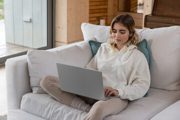 Woman typing on silver laptop while sitting cross-legged on cushioned sofa in wood-paneled cabin © wavebreak3