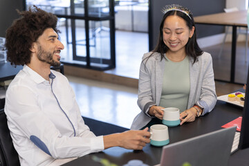 Diverse coworkers holding coffee mugs and cupcakes, wearing birthday headband at desk, laptop