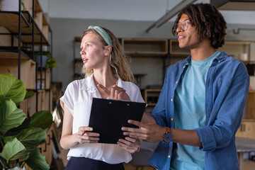 Diverse coworkers pointing at clipboard while discussing plan in shared office near potted plant