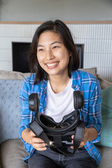 Naklejka premium Asian woman sitting on grey couch in living room by fireplace holding virtual reality headset