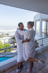 Senior couple wearing white bathrobes and slippers hugging on balcony overlooking pool, ocean
