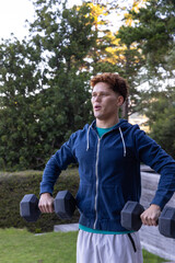 Male in hoodie and shorts performing dumbbell workout in backyard grass with evergreen trees