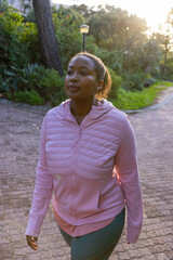 Fototapeta premium African american woman walking in activewear along brick path in park using wireless earbuds
