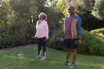Diverse senior couple holding black and purple dumbbells, exercising on lawn in backyard garden