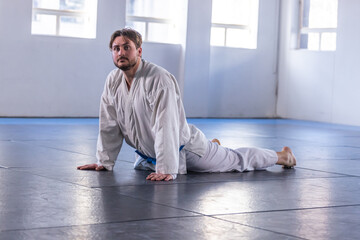 Male martial artist in white gi and blue belt stretching on blue mats in dojo