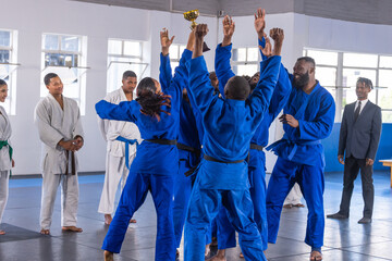 Diverse judo teammates wearing blue gis lifting gold trophy in martial arts hall on mat