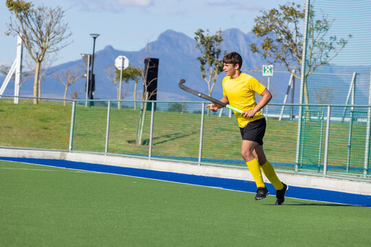 Field hockey player running on bright green turf holding stick next to metal fence and netting