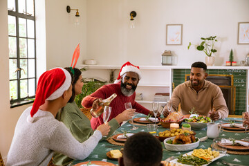 Naklejka premium Diverse family in santa hats at dining table and african american man pouring champagne