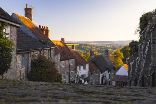 View of quaint cottages with red-tiled roofs cascade down a steep cobbled street, bathed in the warm glow of the setting sun, Shaftesbury, England, United Kingdom.