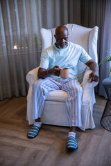 Senior African American man sitting in armchair in living room wearing slippers, holding mug