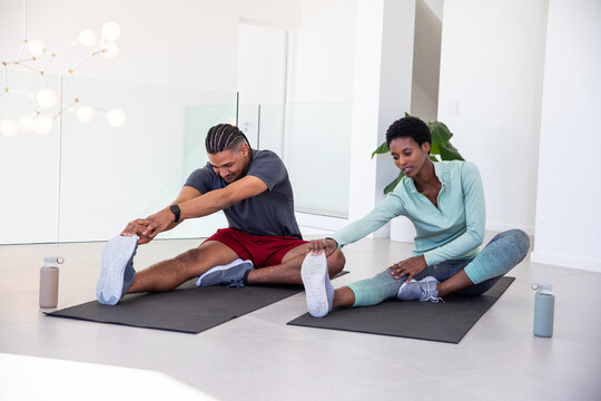 Diverse workout partners stretching hamstrings on black mats in living room with water bottles