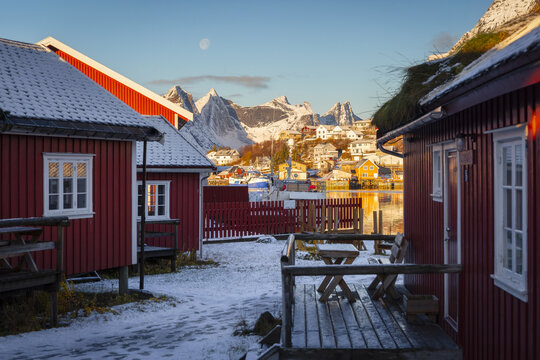 View of rustic red cabins dusted with snow lead the eye towards jagged, snow-capped mountains under a pale sky with a faint moon, Reine, Nordland, Norway.