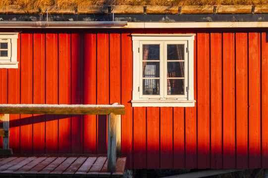 View of a vibrant red wooden building with white-framed windows and a grass-covered roof casting shadows, a picturesque scene in Reine, Nordland, Norway.