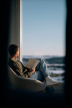 Person Relaxing in Armchair Reading Book by Large Window With City View at Dusk in Cozy Peaceful Home Interior