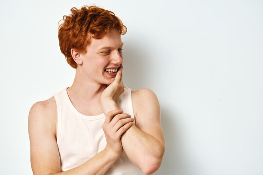 Young man with red hair wearing white tank top laughing and touching face on white background. Casual style, happy expression and natural smile with playful mood in studio.