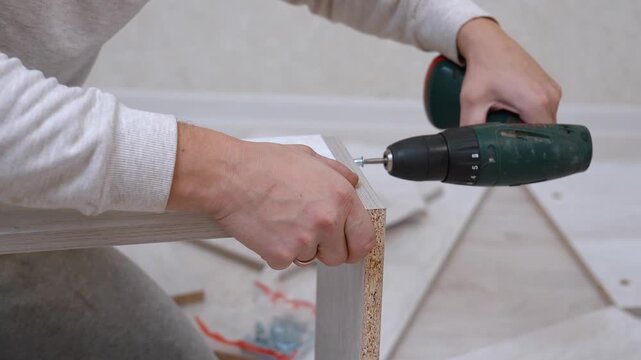 Close-up of hands of man assembling new wooden furniture at home using a cordless screwdriver screwdriver to fasten particle board panels during home renovation by himself