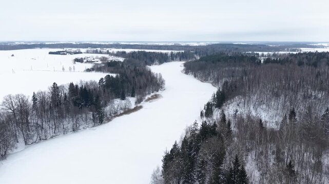 Drone video capturing a serene snowy landscape with a frozen river winding through dense forests. The tranquil winter scene is enveloped in misty, soft daylight, offering a breathtaking view.
