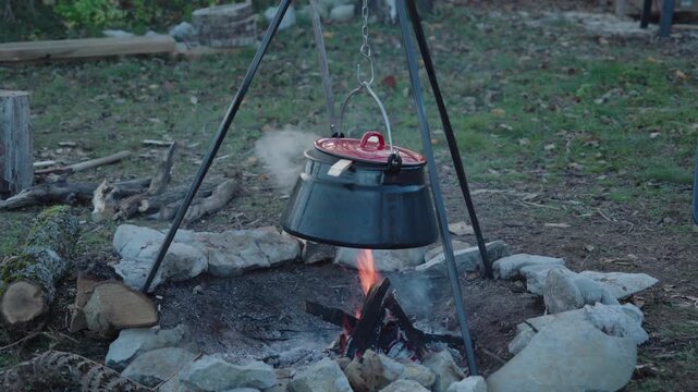 A rustic scene of a black cauldron hanging from a metal tripod over an open campfire. Smoke rises as a meal cooks slowly outdoors.