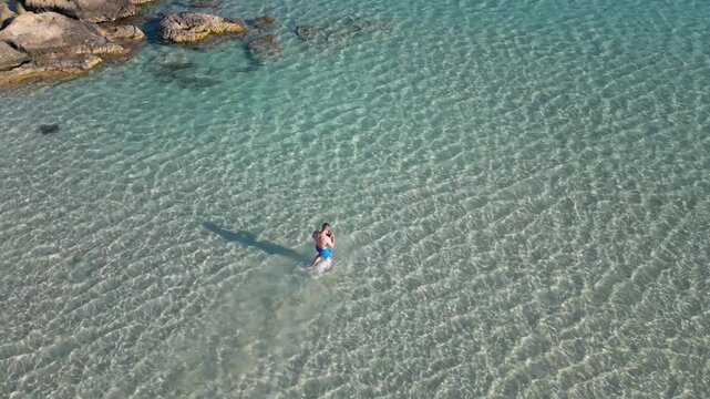 Drone shot: man in trunks walking through shallow turquoise sea water with sun ripples, Elafonisi beach, Crete