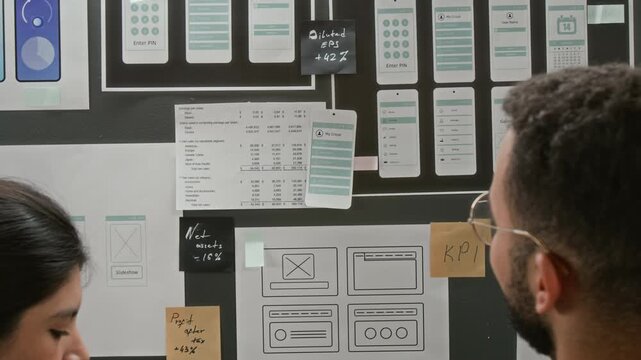 Rear zoom-out shot of two diverse UI designers discussing wireframe concepts on project workflow board, for smartphone operation system or mobile application, brainstorming for usability ideas