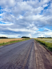 Fototapeta premium Bright rainbow arcs over country road through fields under cloudy sky