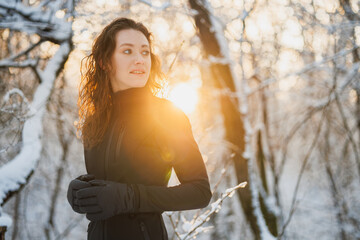 Curly woman in ski suit and gloves looking away in winter forest during sunset 