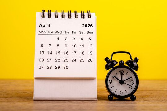 April 2026 desk calendar with a black alarm clock on a wooden table against a yellow background. Concept for time management, planning, and scheduling appointments.