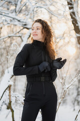 Smiling curly woman in black ski suit and gloves standing in winter forest 