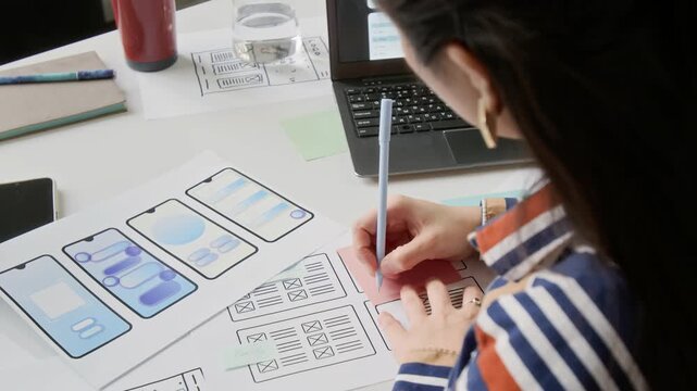 Over-shoulder shot of unrecognizable young female user experience engineer working on new wireframe layouts in office, writing ideas on paper notes, laptop and smartphone on desk nearby
