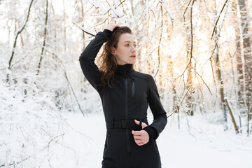Pretty woman in back ski suit standing in winter forest during sunset 