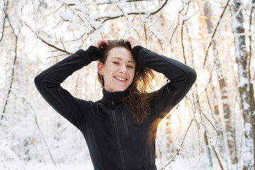 Portrait of cheerful woman in ski suit looking at camera during sunset in winter forest 