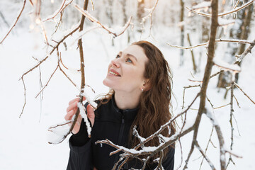 Happy woman in ski suit touching snow on tree in winter forest 