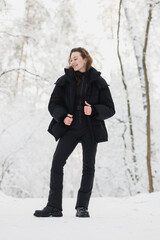 Full length of smiling woman in black outerwear standing in winter forest 