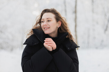 Happy curly woman in black outerwear looking away in winter forest 