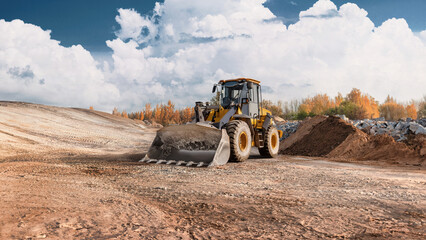 Fototapeta premium Loader is moving dirt on a construction site. The area is preparing for new structures with trees in the background and clouds overhead